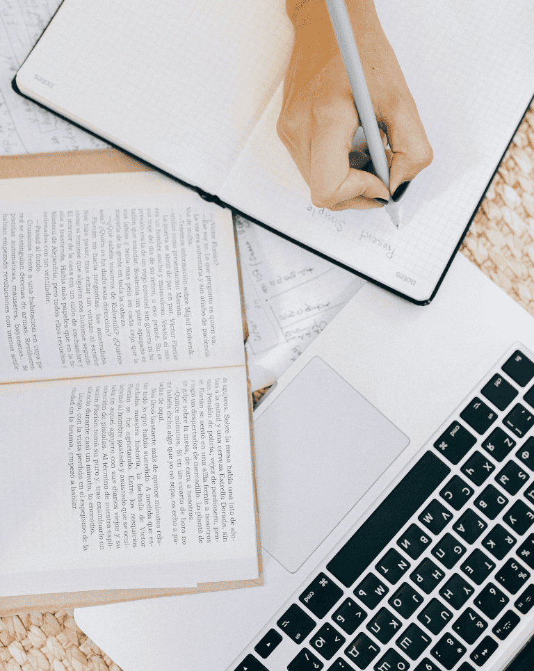 Hand holding pen writing in notebook on top of book and laptop / The Smart Brevity writing style is the best way to write concise, impactful copy.